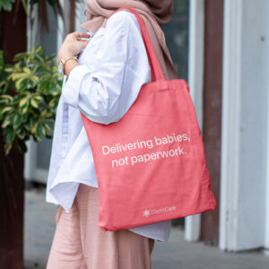 healthtech brand identity Pink ClientCare totebag with the phrase "Delivering babies, not paperwork," held by a woman on a tree-lined street.