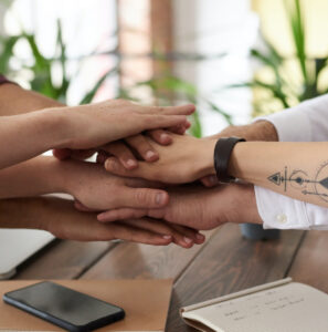 Six hands stacked together in an office setting, symbolizing teamwork and collaboration.