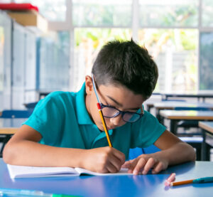 Young student with glasses writing in a notebook inside an empty classroom.