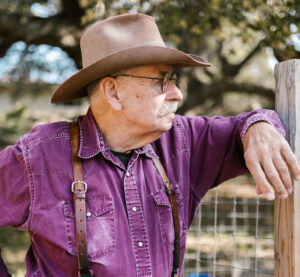 Elderly man with glasses, wearing a hat, leaning on a wooden surface outdoors.