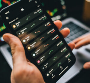 Hand holding a smartphone with cryptocurrency statistics, with a laptop in the background.