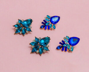 Two pairs of blue and aqua green party earrings on a pink background.