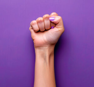 Woman’s fist with purple nails against a purple background, symbolizing feminist solidarity.