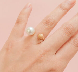 Close-up of a hand wearing a pearl and gold ring on a pink background.