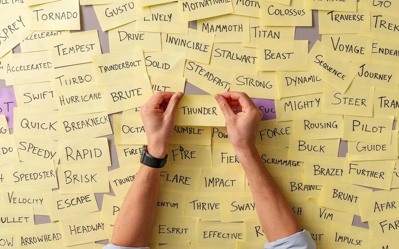 Hands placing a sticky note with a business name idea on a wall filled with naming options for a brand.