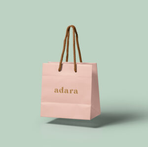 Pink paper bag with adara logo and brown ribbon handle on a green background.