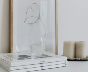 A stack of neutral-toned books with a glass of water, two cylindrical candles, and a framed minimalist floral illustration on a light background.