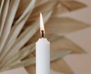 A lit white taper candle with a soft beige background and dried plants, evoking a peaceful ambiance.