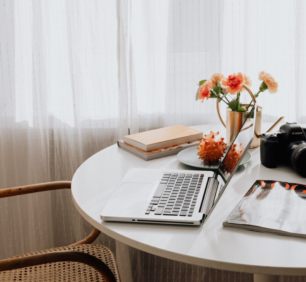 Round desk with laptop, books, flowers, and a camera in natural light.
